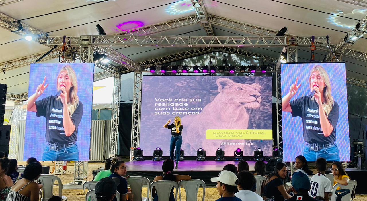 Liza Lopes em palco com telões laterais durante palestra para grande público.